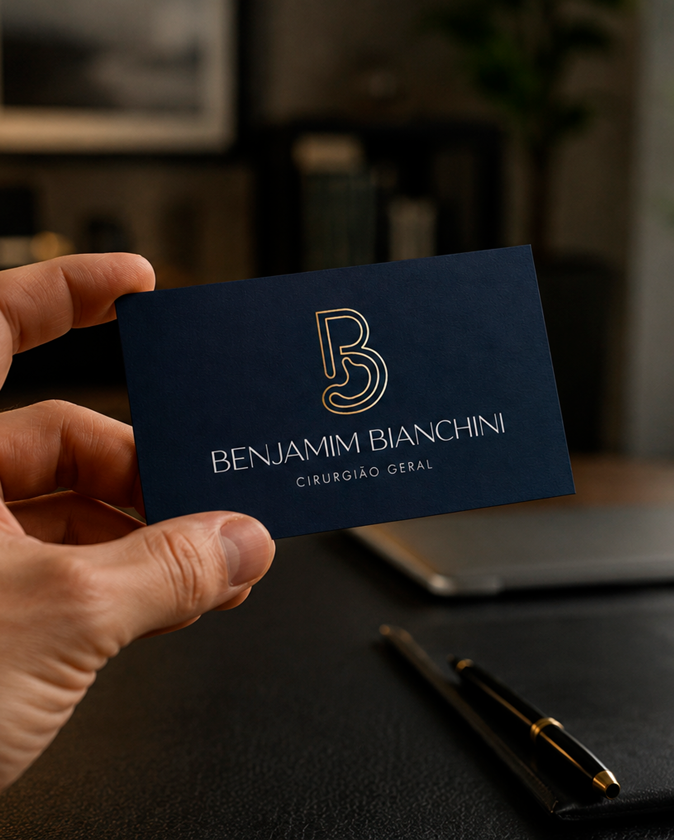 Mão masculina segurando um cartão de visita físico e elegante criado pela Agencia azzy - marketing para medicos para Benjamim Bianchini Cirurgião Geral. O cartão possui fundo azul-marinho fosco com monograma 'BB' em linhas douradas, seguido pelo nome 'BENJAMIM BIANCHINI' e a especialidade 'CIRURGIÃO GERAL'. Ao fundo, vê-se um ambiente de escritório sofisticado com uma caneta sobre a mesa de couro escuro.